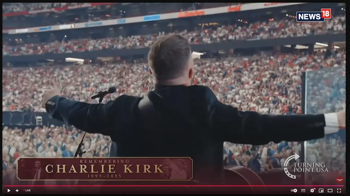 Kirk-3 Auf der Bühne breitet ein Mann seine Arme vor einer riesigen Stadionmenge aus. Auf einer Leinwand ist "Remembering Charlie Kirk 1993-2025" zu lesen, mit den Logos der Sender.