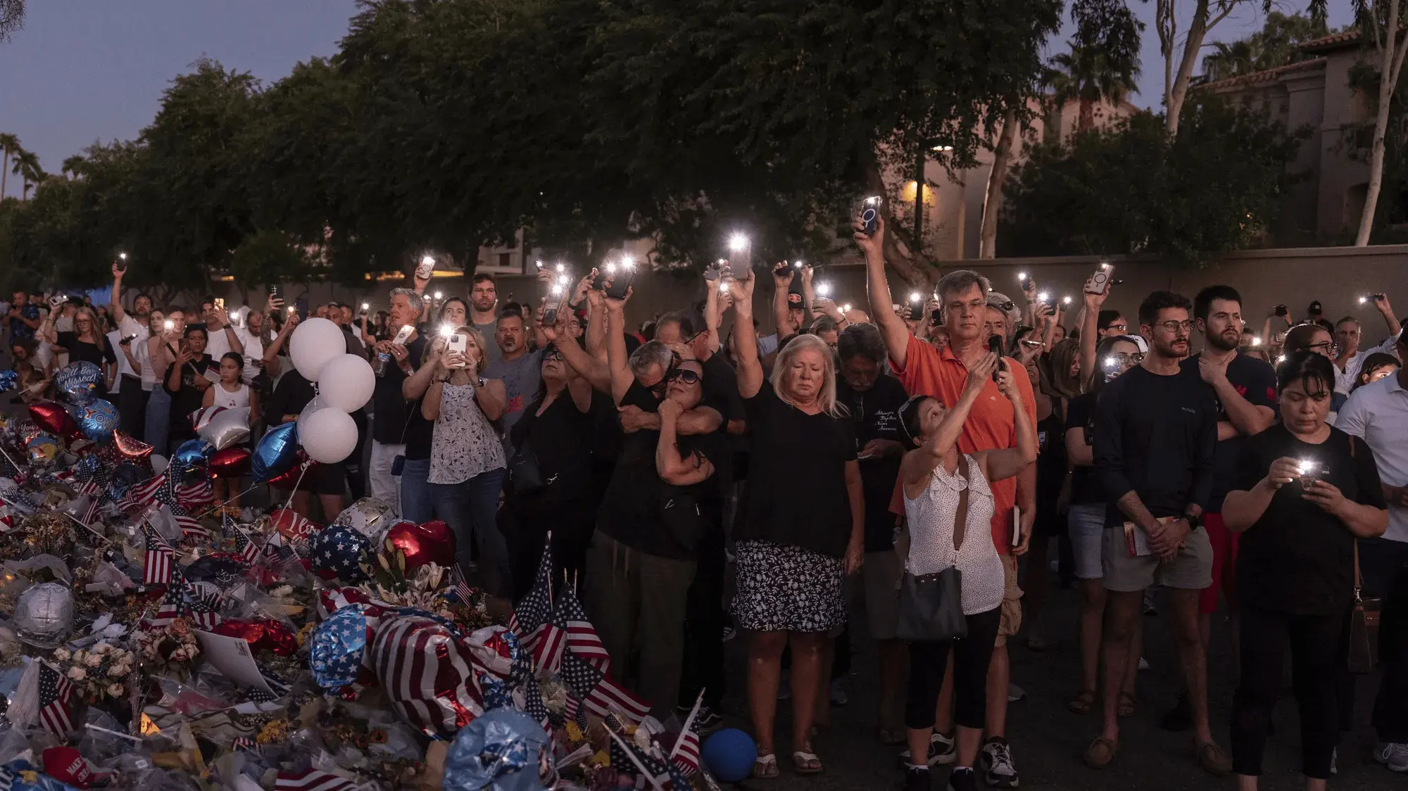 kirk-5 In der Abenddämmerung hebt eine stille Menge leuchtende Telefone vor einem Denkmal aus Ballons, Blumen und Fahnen in die Höhe. Bäume und ein Gebäude verschwinden hinter ihnen.